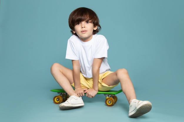 Ein niedlicher Junge der Vorderansicht im weißen T-Shirt und in den gelben Shorts, die grünes Skateboard auf dem blauen Raum reiten