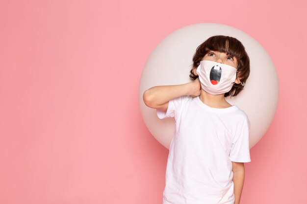 Ein niedlicher Junge der Vorderansicht im weißen T-Shirt und in der lustigen Maske, die weißen Ball auf dem rosa Raum hält