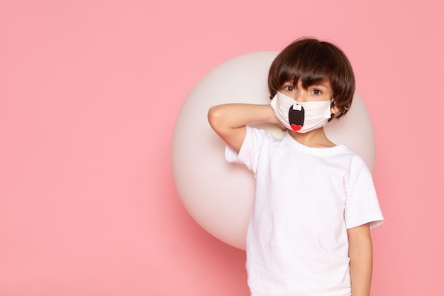 Ein niedlicher Kinderjunge der Vorderansicht im weißen T-Shirt und in der lustigen Maske, die weißen Ball auf dem rosa Schreibtisch hält