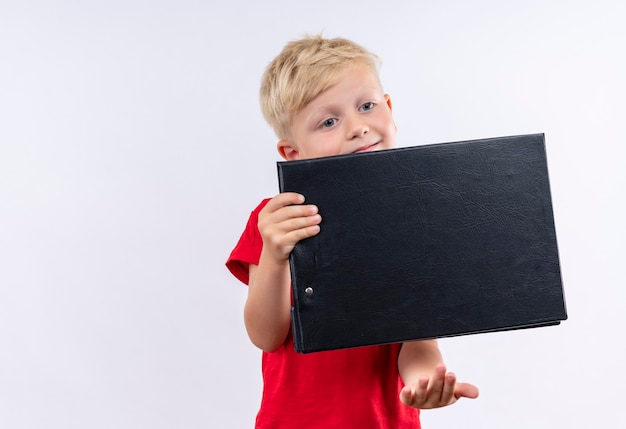 Ein niedlicher kleiner blonder Junge, der rotes T-Shirt trägt, das leeren Ordner auf einer weißen Wand hält