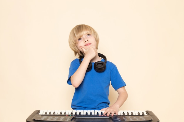 Ein niedlicher kleiner Junge der Vorderansicht im blauen T-Shirt mit schwarzen Kopfhörern, die kleine niedliche Klavier denkende Pose spielen