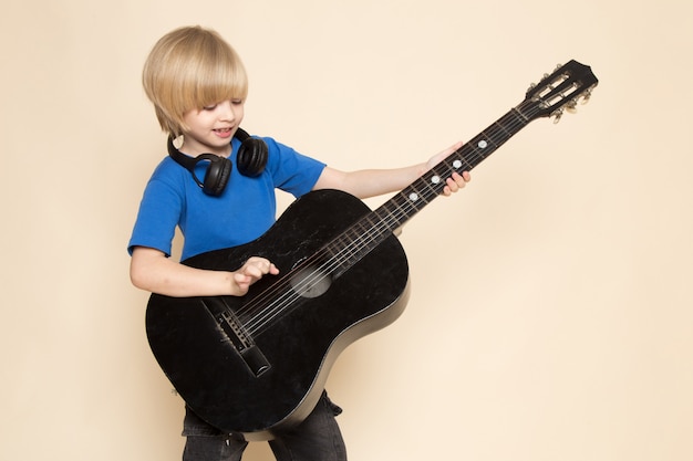 Ein niedlicher kleiner Junge der Vorderansicht im blauen T-Shirt mit schwarzen Kopfhörern, die schwarze Gitarre halten