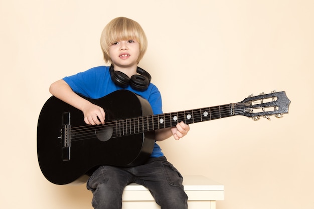 Ein niedlicher kleiner Junge der Vorderansicht im blauen T-Shirt mit schwarzen Kopfhörern, die schwarze Gitarre spielen