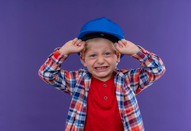 Ein niedlicher kleiner Junge mit blondem Haar, der kariertes Hemd im blauen Helm trägt, der geballte Fäuste erhebt, die auf eine lila Wand schauen