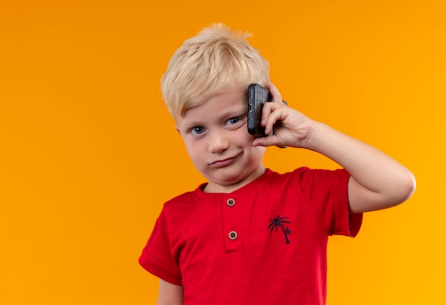 Ein niedlicher kleiner Junge mit blondem Haar trägt rotes Hemd, das Handy hält, während auf einer orange Wand schaut