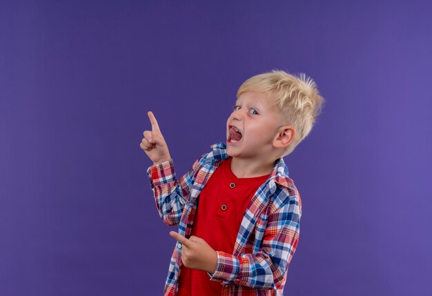 Ein niedlicher kleiner Junge mit den blonden Haaren, die das karierte Hemd tragen, während sie auf eine lila Wand zeigen