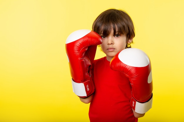 Ein niedliches entzückendes kleines Kind der Vorderansicht entzückend in den blauen Boxhandschuhen und im roten T-Fetzen an der gelben Wand