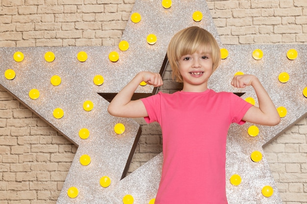 Ein niedliches kleines Kind der Vorderansicht in den grauen Jeans des rosa T-Shirts auf dem Stern gestaltete gelben Stand und hellen Hintergrund