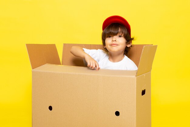 Ein niedliches kleines Kind der Vorderansicht in der roten Kappe des weißen T-Shirts innerhalb der braunen Box auf der gelben Wand