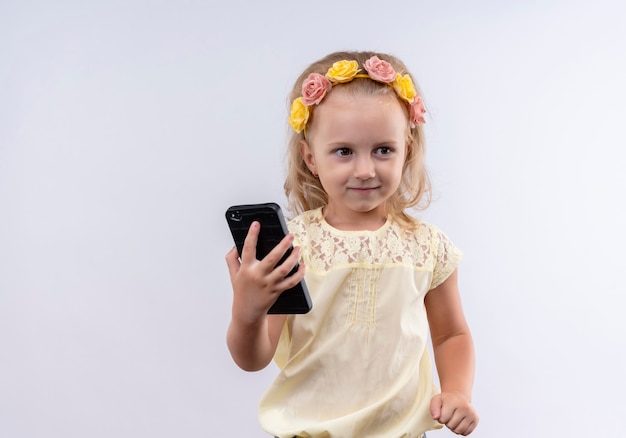 Ein niedliches kleines Mädchen, das gelbes Hemd im Blumenstirnband hält, hält ein Handy, während Seite auf einer weißen Wand schaut