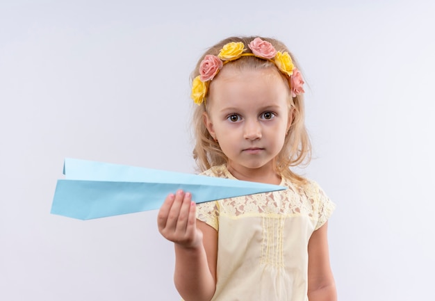 Ein niedliches kleines Mädchen, das gelbes Hemd im fliegenden Papierflugzeug des floralen Stirnbandes trägt, während auf einer weißen Wand schaut