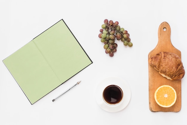 Ein offenes Notizbuch; Stift; Trauben; Kaffee; Croissant und halbierte Orange auf weißem Hintergrund