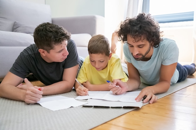 Ein paar homosexuelle Eltern helfen fokussierten Jungen bei der Hausaufgabe in der Schule, liegen zu Hause auf dem Boden, schreiben oder zeichnen in Papieren. Konzept der Familie und der schwulen Eltern