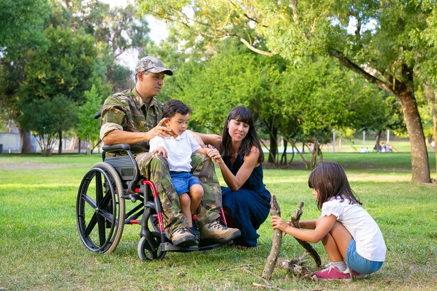 Ein paar nachdenkliche und friedliche Eltern verbringen ihre Freizeit mit Kindern im Freien und arrangieren Brennholz für Feuer auf Gras. Behinderte Veteran oder Familie im Freien Konzept