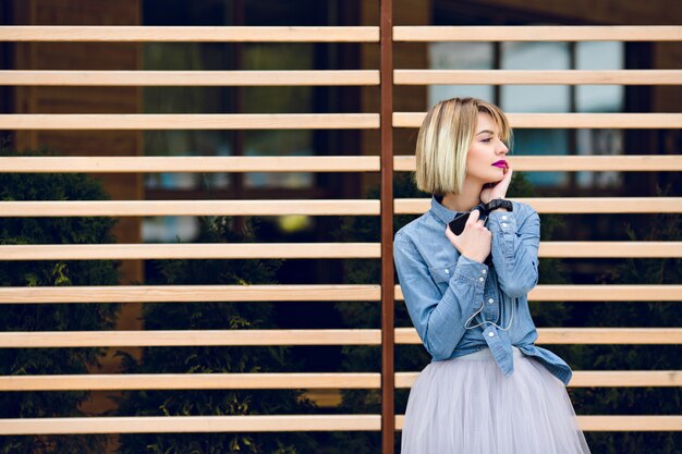 Ein Porträt eines verträumten blonden Mädchens mit leuchtend rosa Lippen und nacktem Make-up, das Musik auf einem Smartphone mit gestreiften Holzbalken hinter sich hört