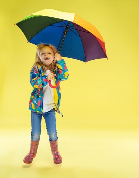 Ein Porträt in voller Länge eines hellen modischen Mädchens in einem Regenmantel, der einen Regenschirm der Regenbogenfarben auf gelber Studiowand hält