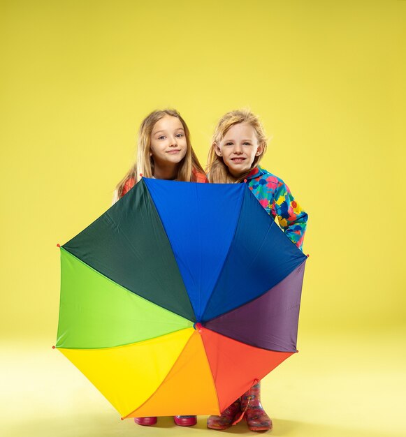 Ein Porträt in voller Länge eines hellen modischen Mädchens in einem Regenmantel, der einen Regenschirm der Regenbogenfarben auf gelber Studiowand hält