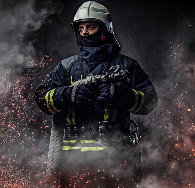 Ein professioneller feuerwehrmann in uniform hält den feuerwehrschlauch in feuerfunken und rauch auf dunklem hintergrund.