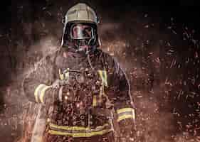 Kostenloses Foto ein professioneller feuerwehrmann in uniform und eine sauerstoffmaske, die in feuerfunken und rauch auf dunklem hintergrund steht.