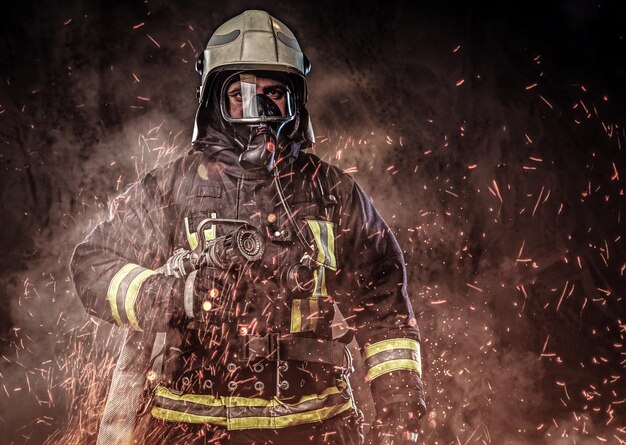 Ein professioneller Feuerwehrmann in Uniform und eine Sauerstoffmaske, die in Feuerfunken und Rauch auf dunklem Hintergrund steht.