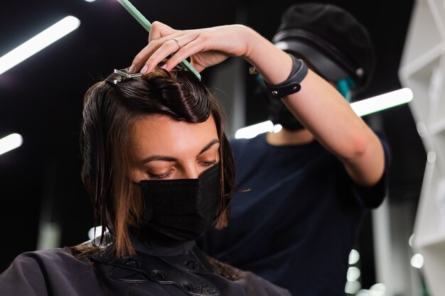 Ein professioneller Friseur macht einen Kundenhaarschnitt. Das Mädchen sitzt in einer Maske in Schönheit im Salon