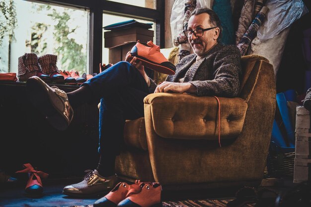 Ein reifer Schuhdesigner sitzt auf dem Sessel seiner Werkstatt und hält einen Schuh aus seiner letzten Kollektion in der Hand.