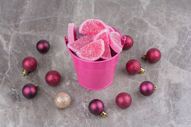 Ein rosa Eimer voller süßer Marmeladen mit roten Weihnachtskugeln auf Marmorhintergrund.