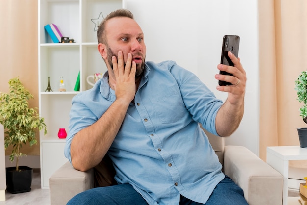 Ein schockierter erwachsener slawischer Mann sitzt auf einem Sessel und legt die Hand auf das Gesicht, während er das Telefon im Wohnzimmer betrachtet