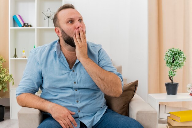 Ein schockierter erwachsener slawischer Mann sitzt auf einem Sessel und legt die Hand auf den Mund, wobei er die Seite im Wohnzimmer betrachtet