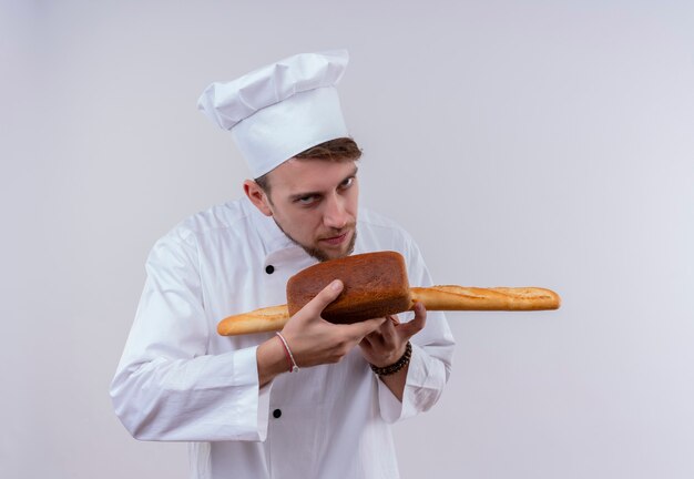 Ein schöner junger bärtiger Kochmann, der weiße Kochuniform und Hut trägt, der Baguettebrot mit Laibbrot riecht, während er auf eine weiße Wand schaut