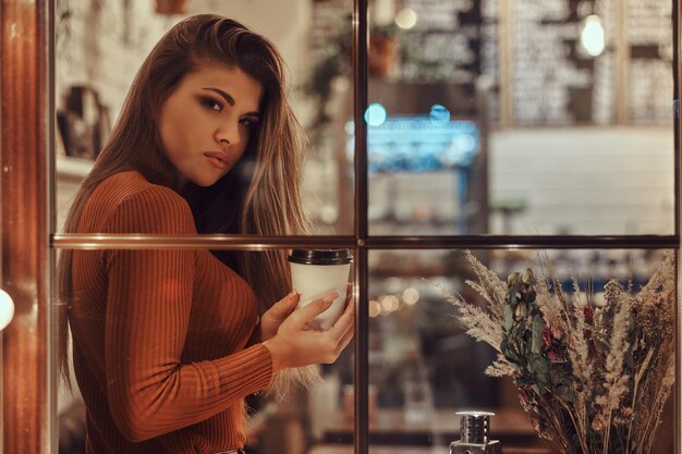 Ein schönes Mädchen mit Pullover, das einen Kaffee hält, während es auf einem Fensterbrett im Café sitzt