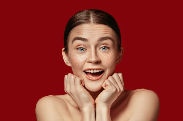 Ein schönes weibliches Gesicht. Eine perfekte und saubere Haut der jungen überraschten kaukasischen Frau auf rotem Studiohintergrund.