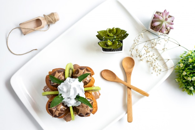 Ein Schoko-Dessert von oben mit geschnittenen Kiwis und Vanillepudding im weißen Schreibtisch zusammen mit Holzlöffeln und Pflanzen auf dem weißen Tischfrucht exotisch süß