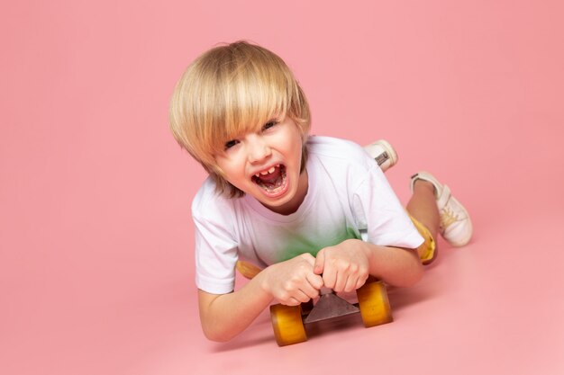 Ein schreiender blonder Junge der Vorderansicht, der grünes Skateboard auf dem rosa Boden reitet