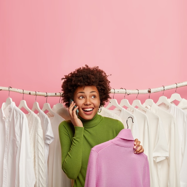 Ein Schuss einer glücklichen Afroamerikanerin wählt neue Kleidung im Ausstellungsraum aus, nimmt einen lila Rollkragenpullover an den Kleiderbügeln auf und steht über einem Satz weißer Kleidung an den Kleiderbügeln