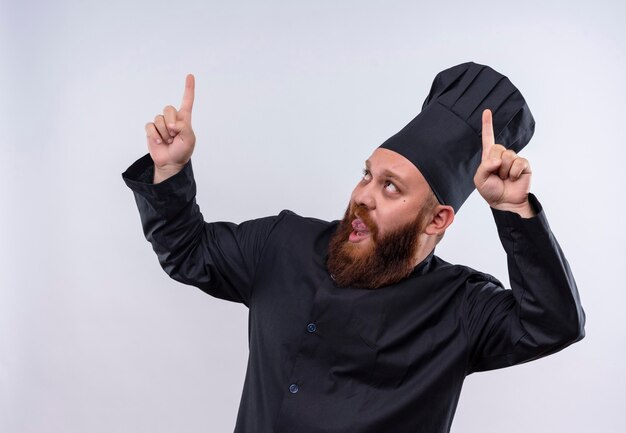 Ein selbstbewusster bärtiger Kochmann in der schwarzen Uniform, die mit Zeigefingern auf einer weißen Wand zeigt