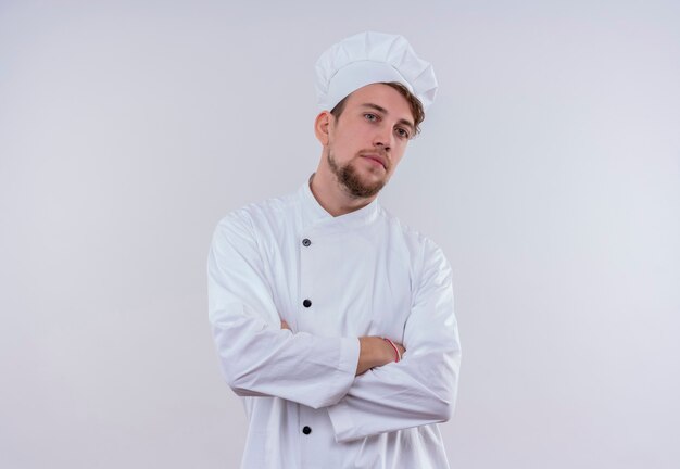 Ein selbstbewusster hübscher junger bärtiger Kochmann, der weiße Kochuniform und Hut hält Hände gefaltet, während er auf eine weiße Wand schaut
