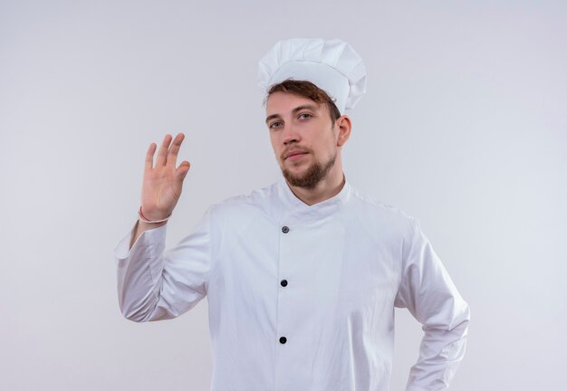 Ein selbstbewusster hübscher junger bärtiger Kochmann, der weiße Kochuniform und Hut trägt und leckere ok Geste mit den Fingern auf einer weißen Wand zeigt