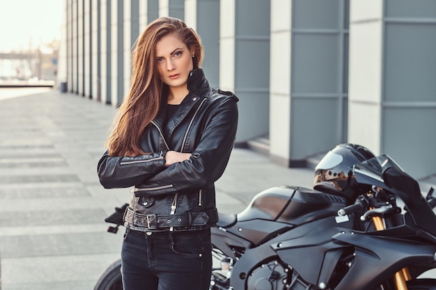 Ein selbstbewusstes Biker-Mädchen, das eine schwarze Lederjacke mit verschränkten Armen neben ihrem Superbike vor einem Gebäude trägt.