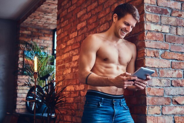 Ein sexy, tätowierter, hemdloser Mann mit Touchpad lehnt an der Ziegelwand. Lächelnd und wegschauend.