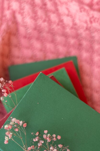 Ein Stapel roter und grüner Bücher mit trockenen Blumen auf einem rosa warmen Strickpullover