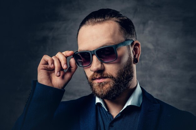 Ein stilvoller bärtiger Mann in Anzug und Sonnenbrille auf dunkelgrauem Hintergrund.