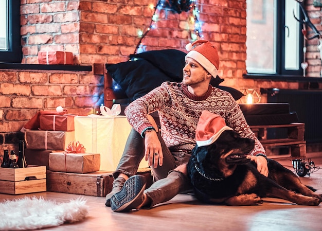 Ein stilvoller junger Mann, der zur Weihnachtszeit mit seinem reinrassigen Rottweiler in einem dekorierten Wohnzimmer sitzt.