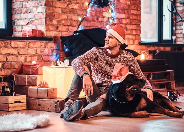Ein stilvoller junger Mann, der zur Weihnachtszeit mit seinem reinrassigen Rottweiler in einem dekorierten Wohnzimmer sitzt.