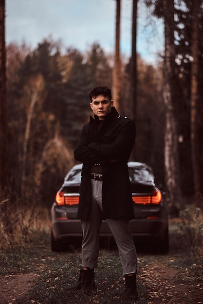 Ein stilvoller junger mann in einem schwarzen mantel mit verschränkten armen steht abends neben einem luxusauto im herbstwald.