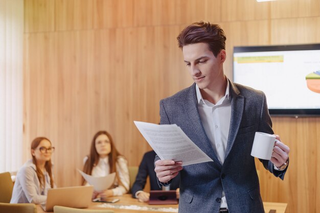 Ein stilvoller Mann in einer Jacke und einem Hemd mit einer Tasse Kaffee in der Hand steht und liest Dokumente vor dem Hintergrund der Arbeitskollegen im Büro