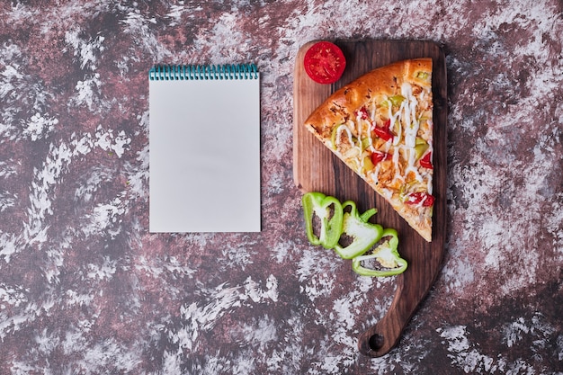 Kostenloses Foto ein stück pizza auf einem holzbrett mit einem rezeptbuch auf dem marmor.