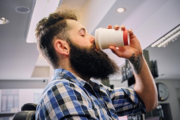 Ein stylischer bärtiger Hipster-Mann in einem Fleece-Shirt trinkt Kaffee in einem Friseursalon.