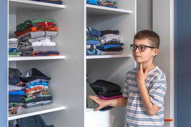 Ein süßer Junge mit Brille steht in der Nähe eines Kleiderschranks und überlegt, was er anziehen soll.