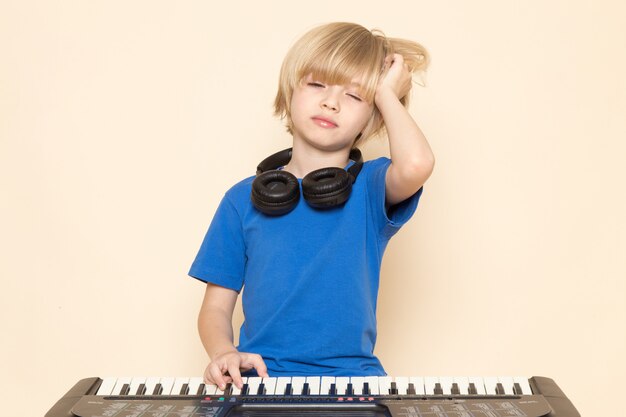 Ein süßer kleiner Junge der Vorderansicht im blauen T-Shirt mit schwarzen Kopfhörern, die kleines niedliches Klavier spielen, das Schlaf will